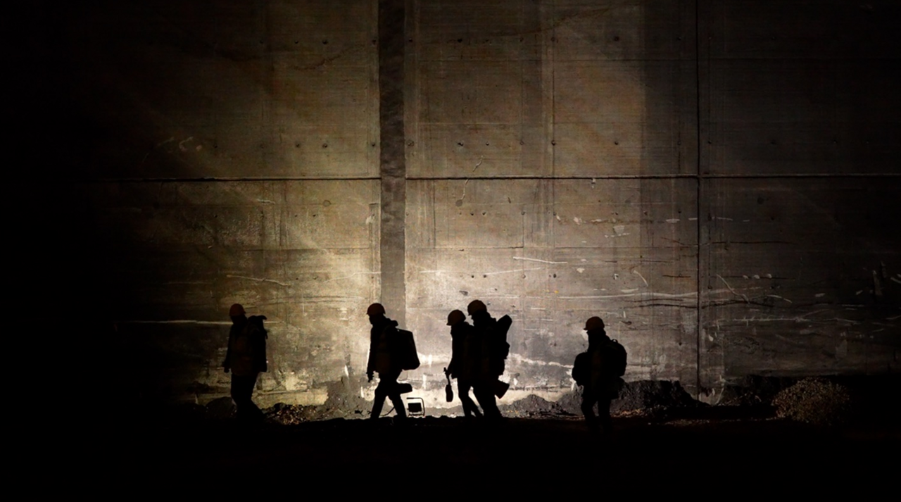 Eine schattenhafte Gruppe von fünf Menschen mit Bauhelmen, dicken Jacken, Rucksäcken bewegt sich im Scheinwerferlicht an einer dunklen Mauer entlang